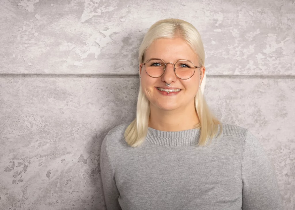 Eine Person mit blonden Haaren und runder Brille lächelt vor einer grauen Wand.