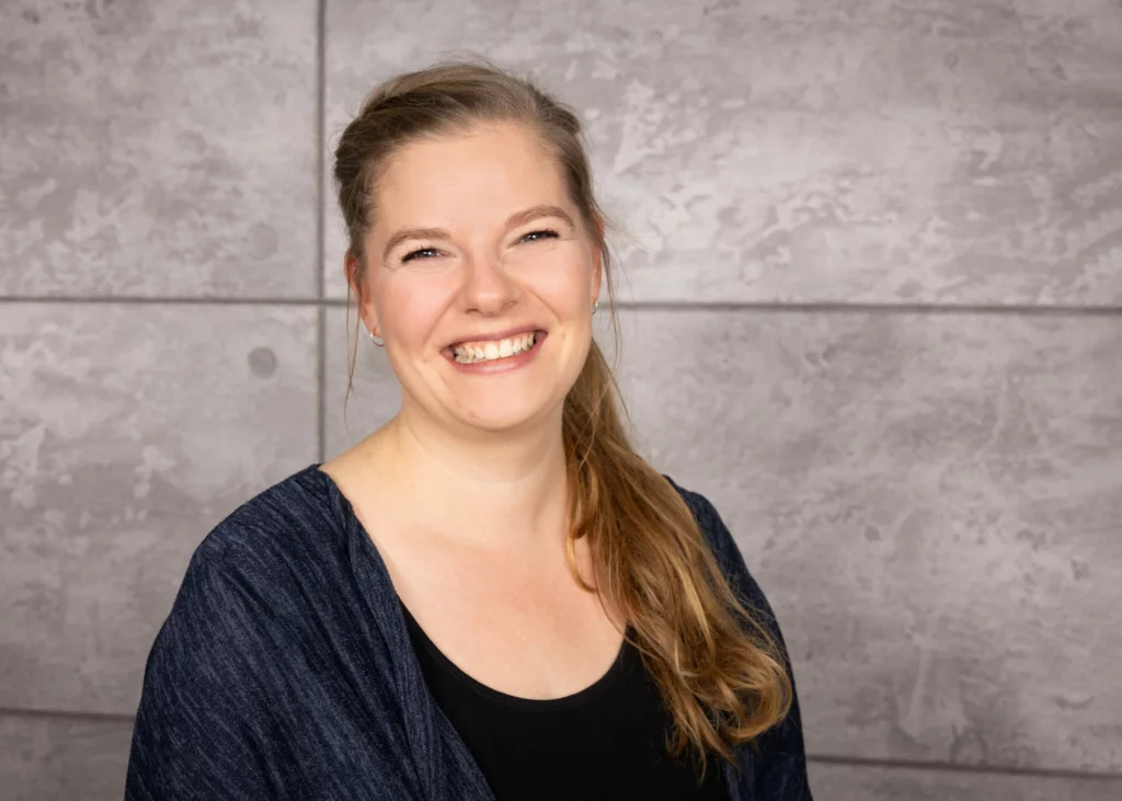 Lächelnde Frau mit langen, blonden Haaren und einem blauen Oberteil vor einer grauen Wand.