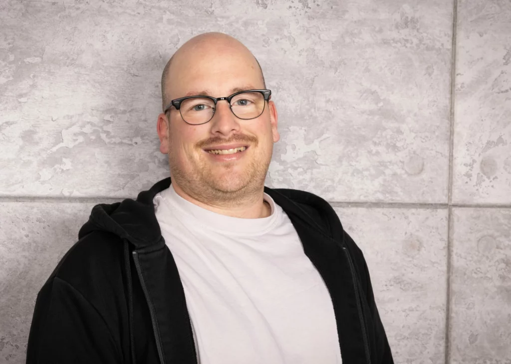 Mann mit Glatze und Brille, der in die Kamera lächelt, vor einer grauen Wand.