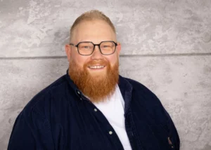 Mann mit braunem Bart und Brille, der in die Kamera lächelt, vor einer grauen Wand.