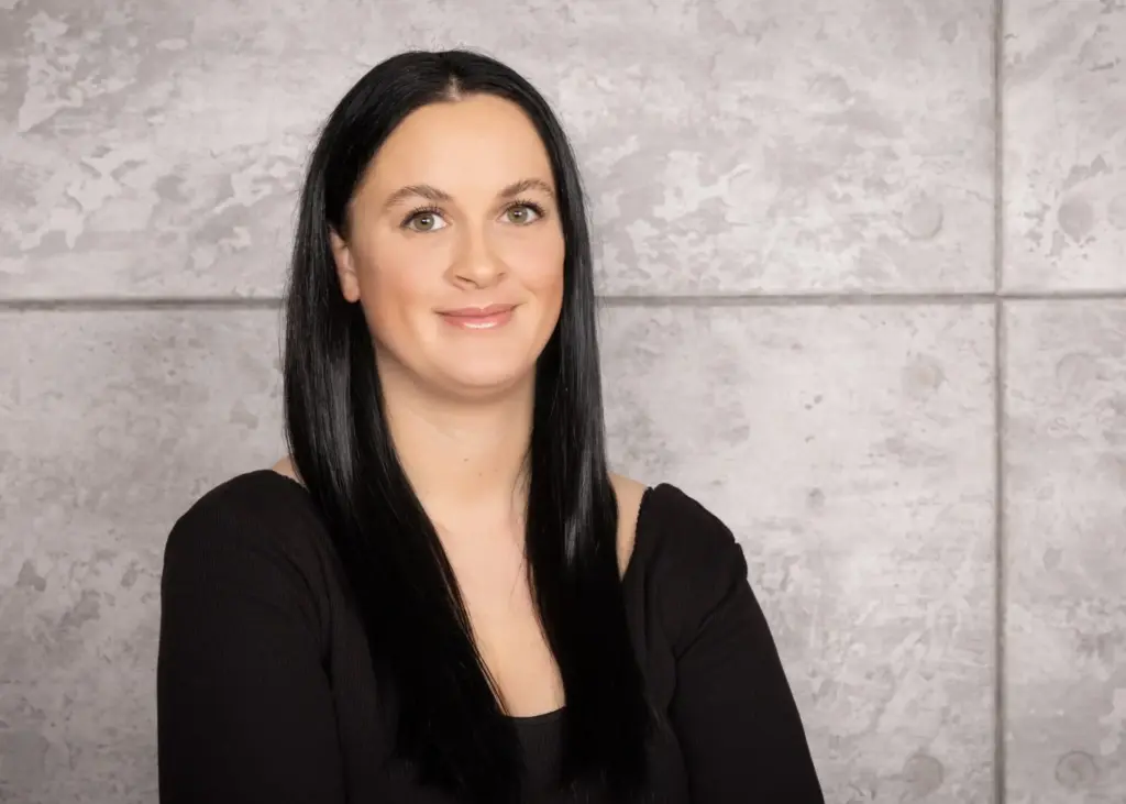 Porträt einer Frau mit langen schwarzen Haaren, die in einem schwarzen Oberteil vor einer grauen Wand steht.