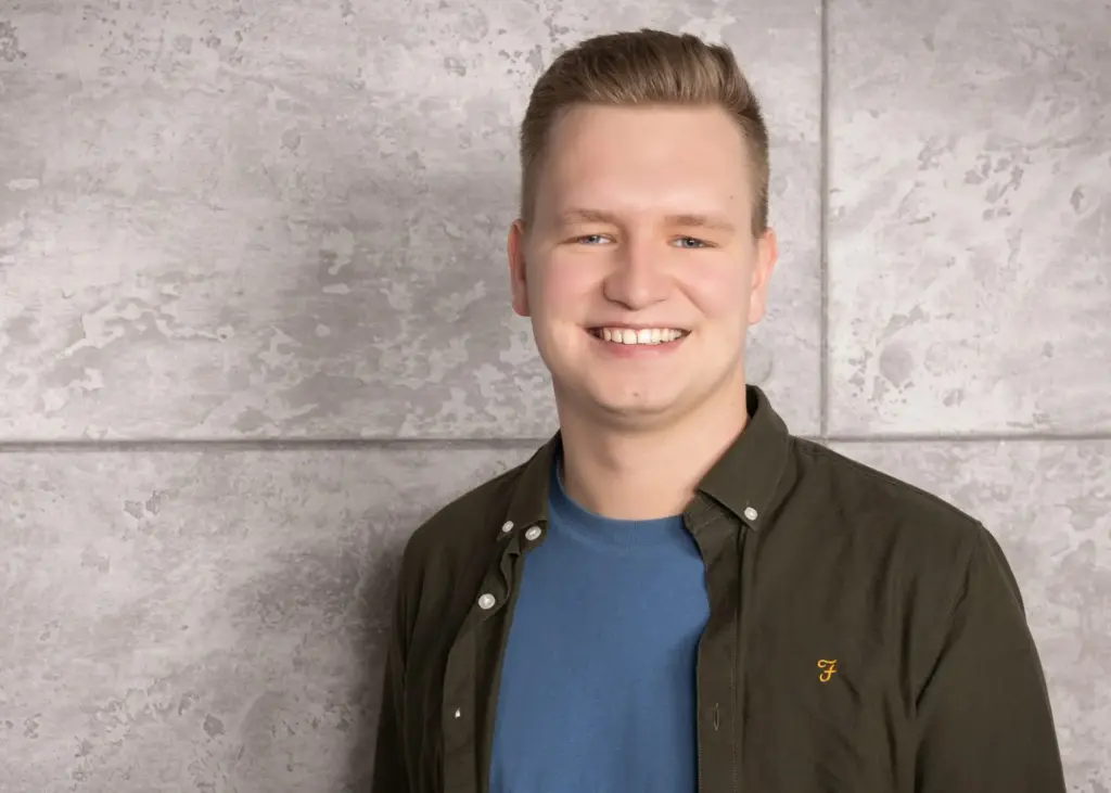Ein junger Mann mit kurzem, blondem Haar und einem lächelnden Gesicht, trägt ein dunkelgrünes Hemd und ein blaues T-Shirt.