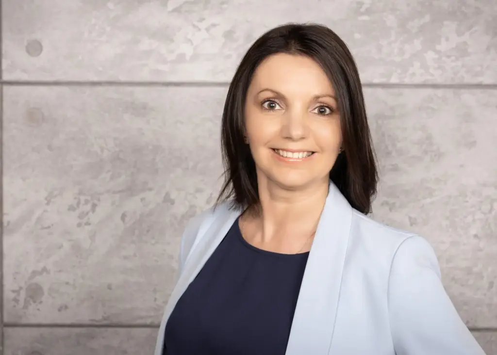 Frau mit braunen Haaren in blauer Bluse und Jacke vor einer grauen Wand.
