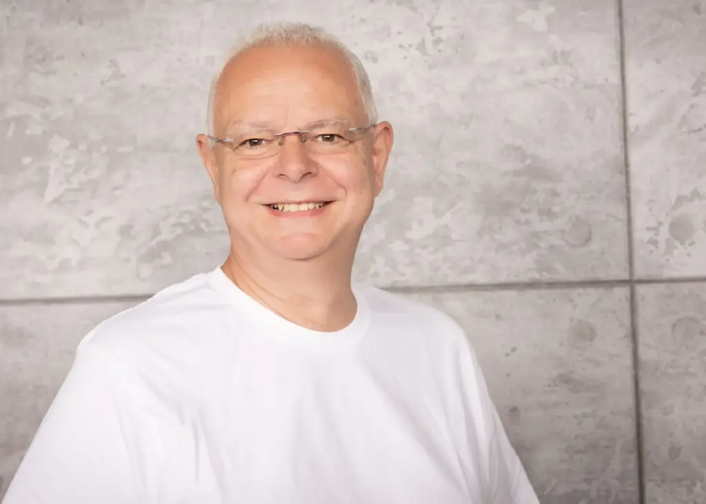 Ein Mann mit Brille und weißem T-Shirt lächelt vor einer grauen Wand.