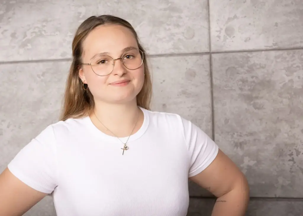 Junge Frau mit Brille und langen Haaren in einem weißen T-Shirt vor einer grauen Wand.