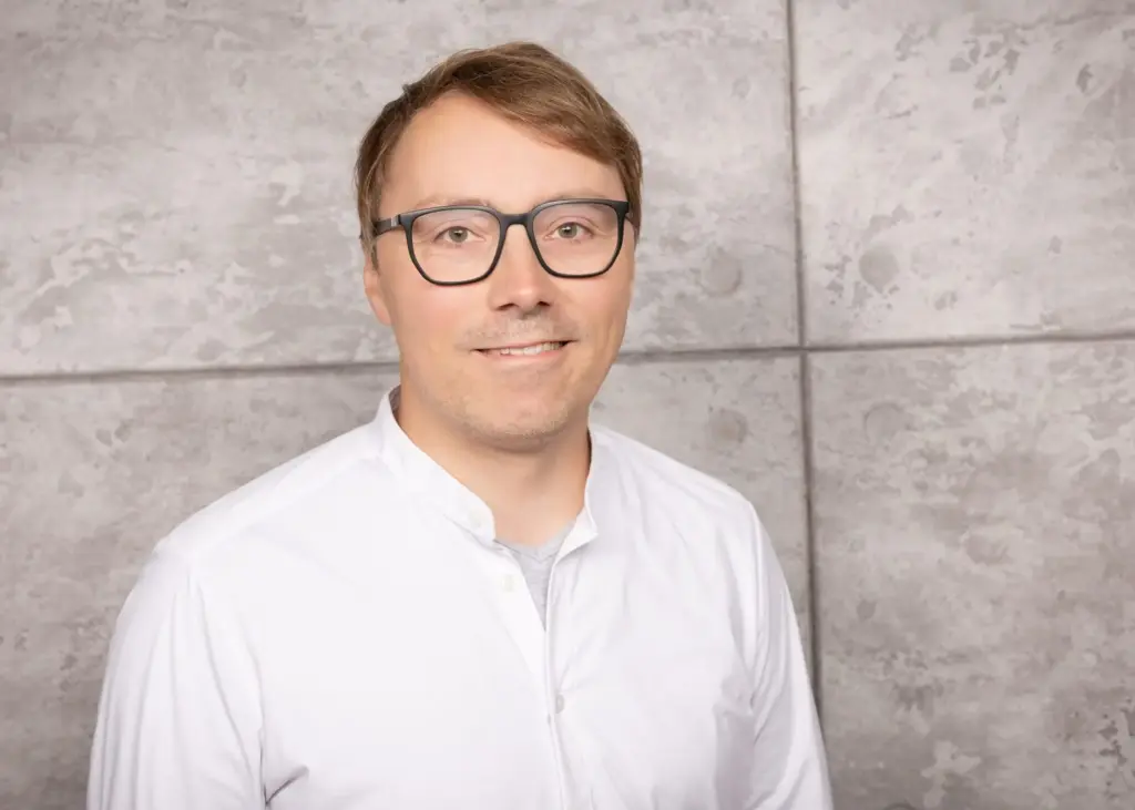 Mann mit Brille und kurzem, braunem Haar in einem weißen Hemd vor einer grauen Wand.