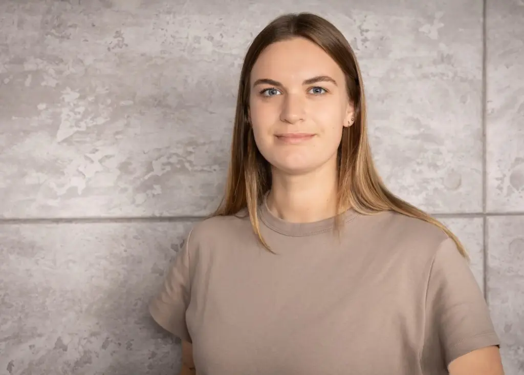 Junge Frau mit blonden Haaren und beigem T-Shirt vor einer grauen Wand.