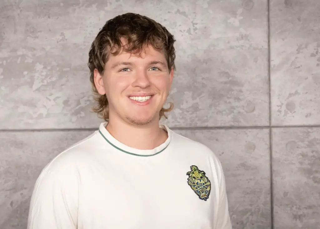 Junger Mann mit lockigem Haar und einem weißen T-Shirt, das ein Logo zeigt, lächelt in die Kamera.