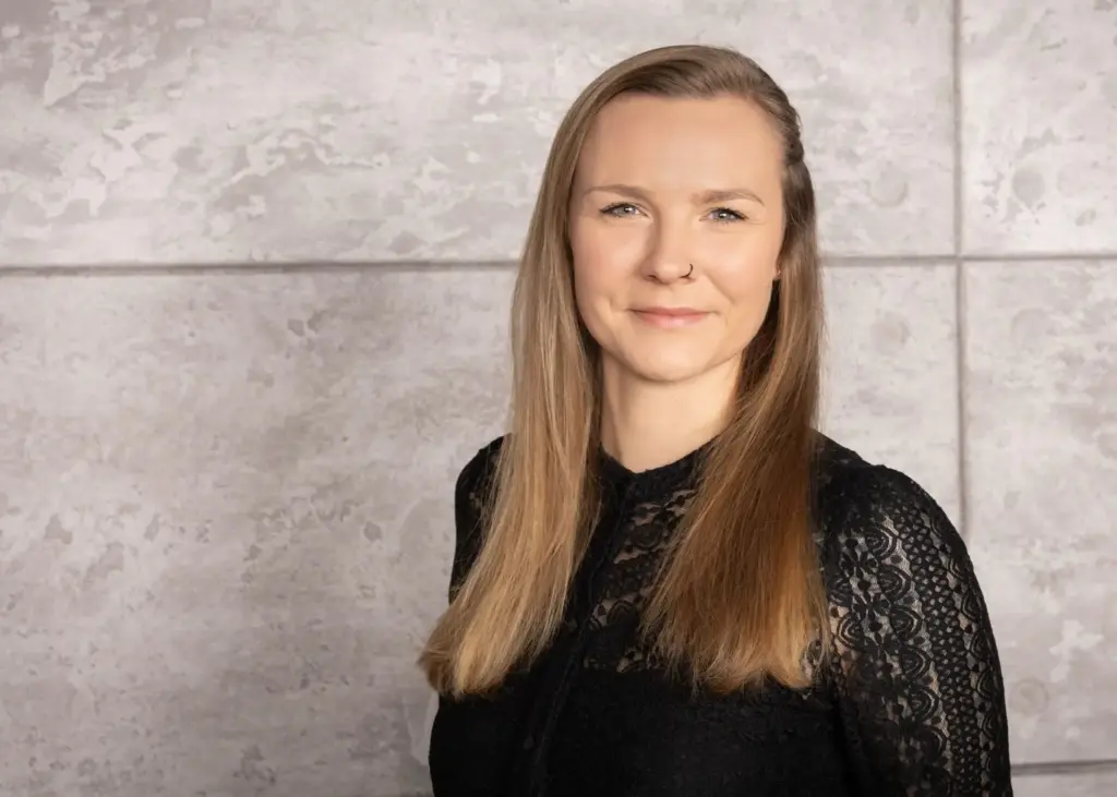 Frau mit langen, blonden Haaren und schwarzem Oberteil vor einer grauen Wand.