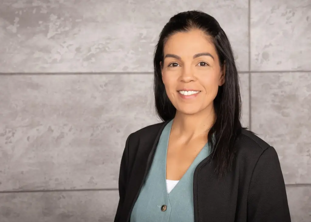 Frau mit langen schwarzen Haaren, die einen schwarzen Blazer und ein hellblaues Oberteil trägt, lächelt in die Kamera.