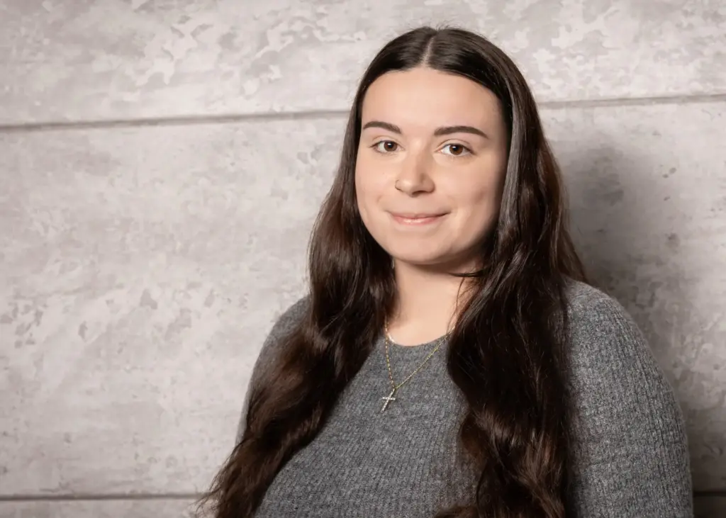 Junge Frau mit langen, dunklen Haaren, die in einem grauen Pullover vor einer neutralen Wand steht.