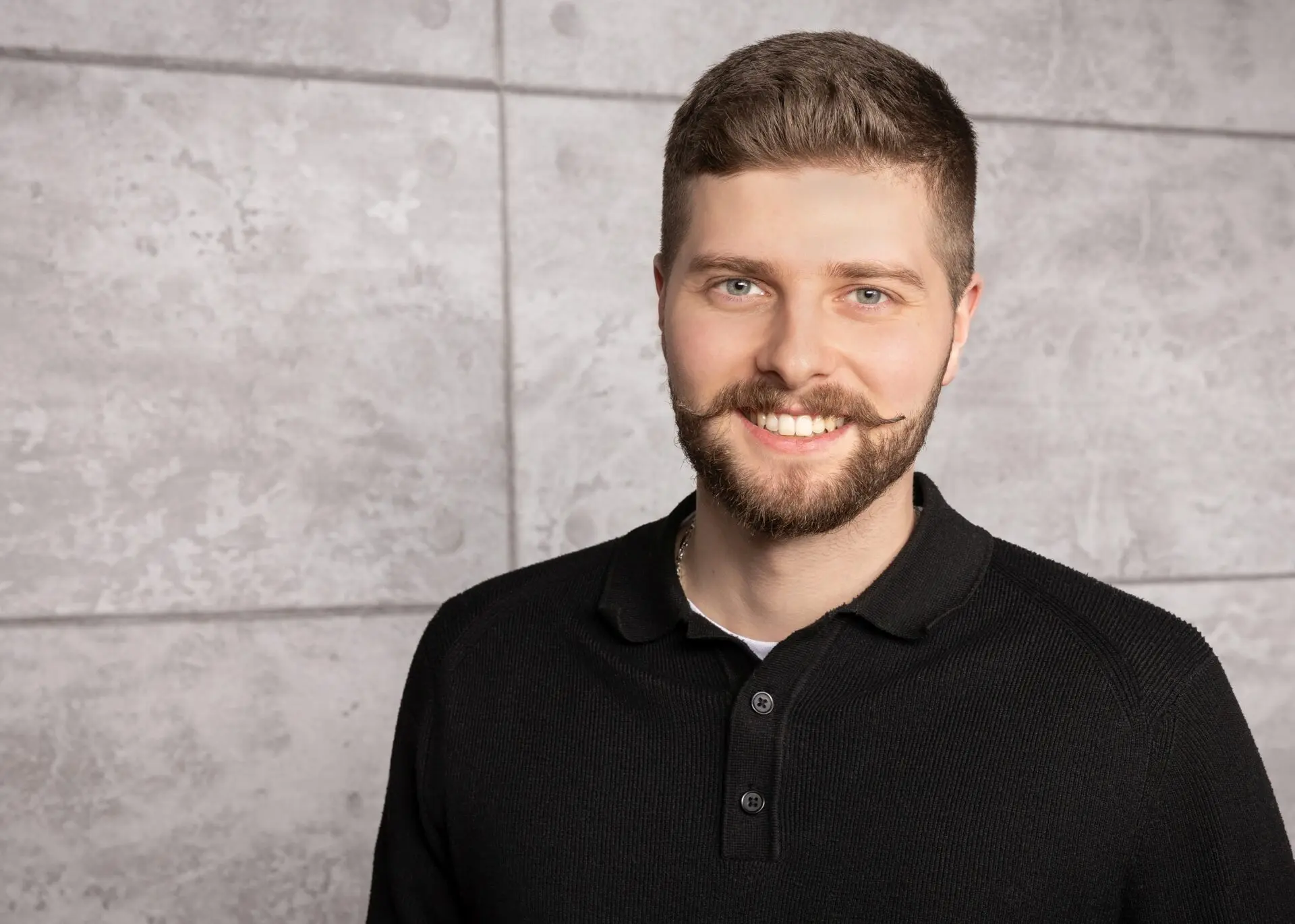 Mann mit Bart und lächelndem Gesicht, trägt ein schwarzes Poloshirt vor einer grauen Wand.