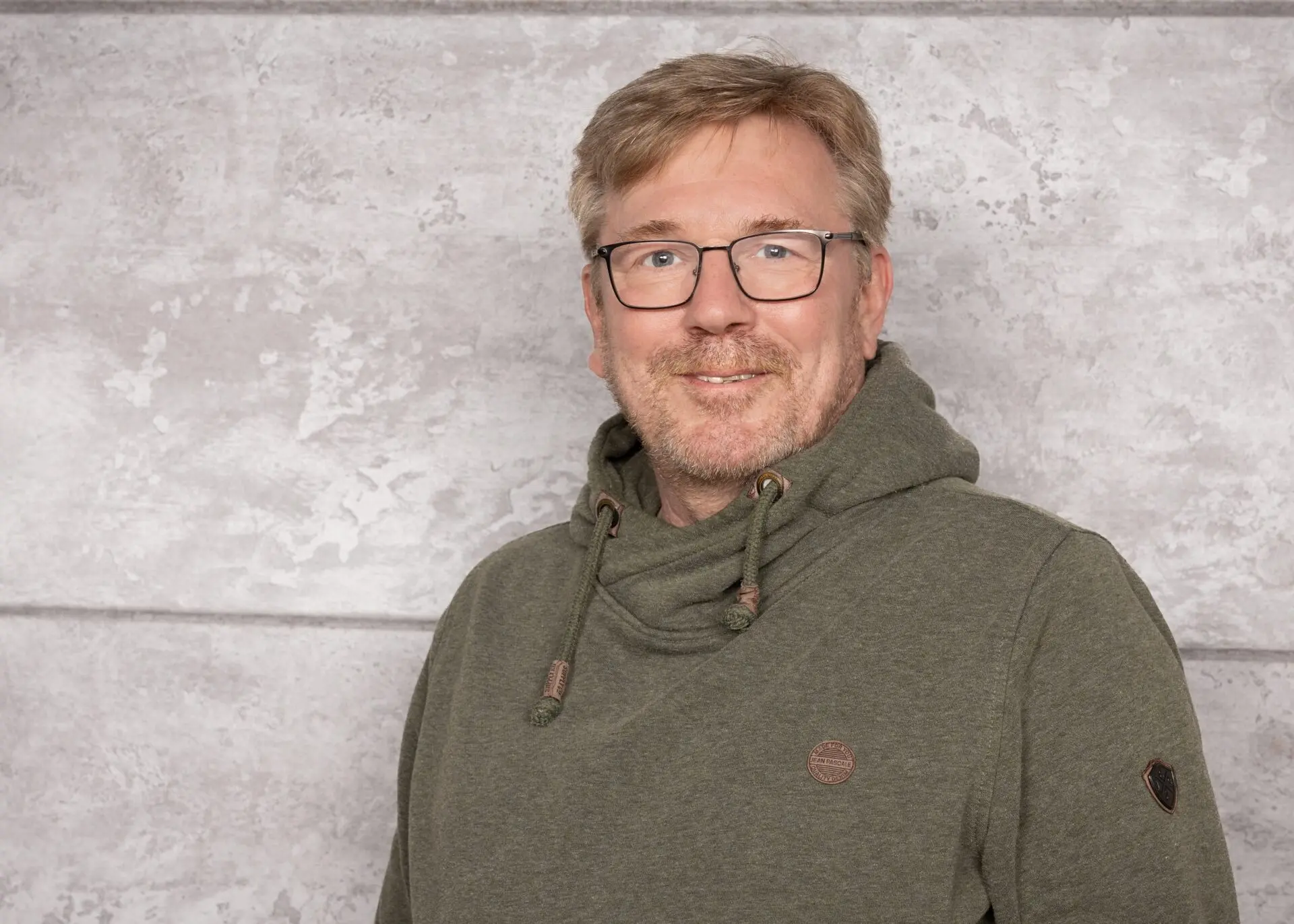 Mann mit Brille und grünem Hoodie, der vor einer grauen Wand steht und lächelt.