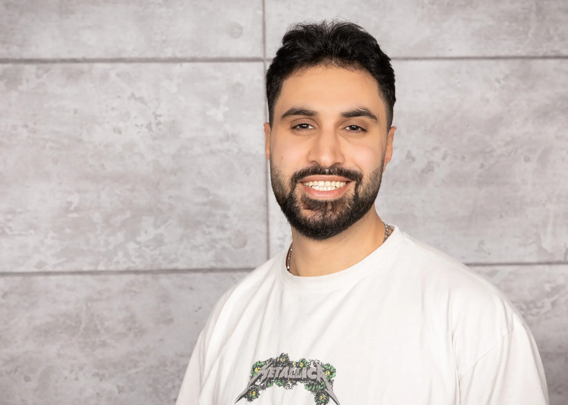Ahmad Ali Ramadan Barazi, Pädagogische Fachkraft, mit dunkelbraunen Haaren und Bart, trägt ein helles T-Shirt und lächelt vor einem grau geprägten Hintergrund.