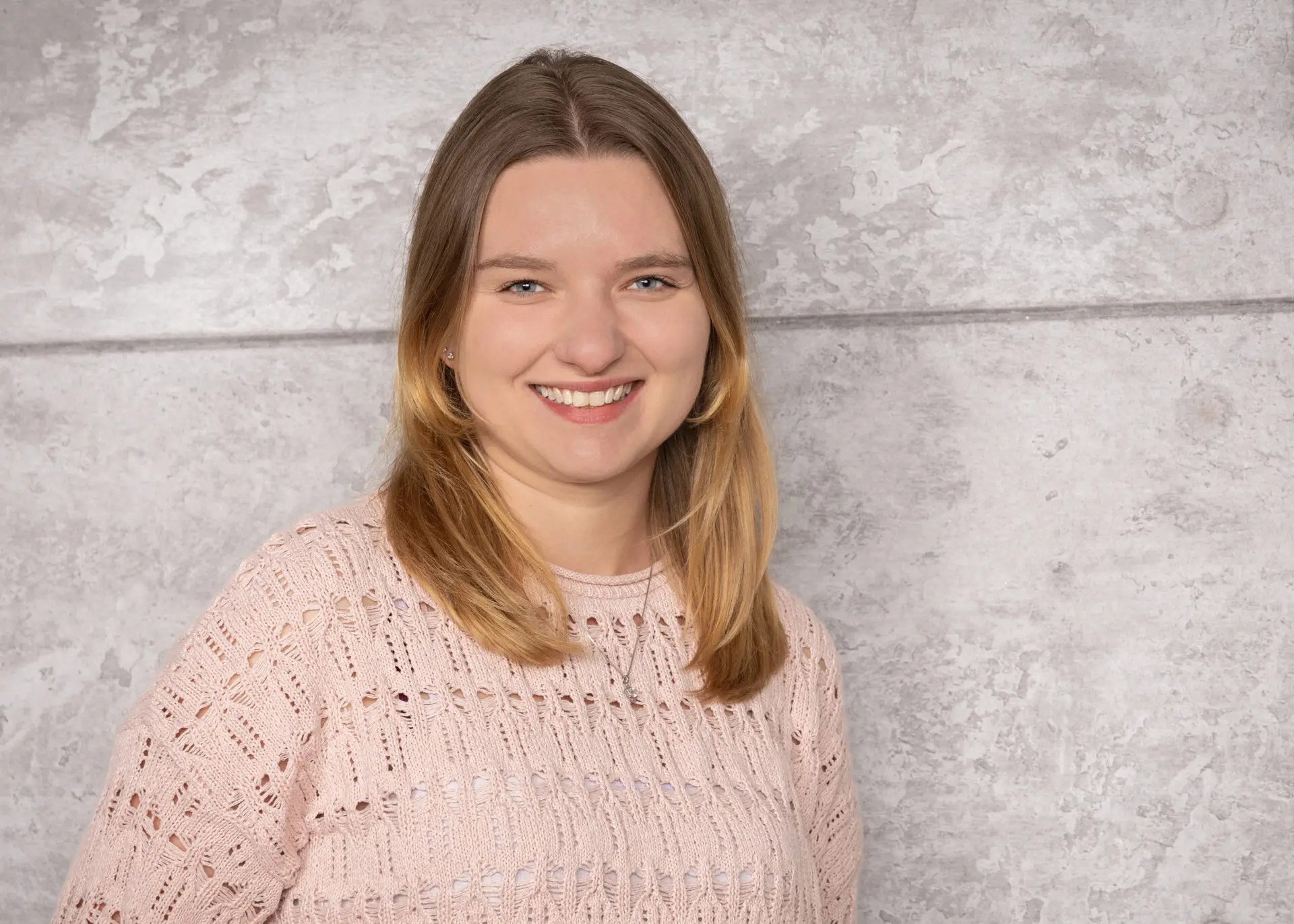 Vivien Kneer lächelt freundlich vor grauem Hintergrund, mit schulterlangem hellbraunem Haar und hellrosa Strickpullover mit Lochmuster, klarer Blick nach vorn.